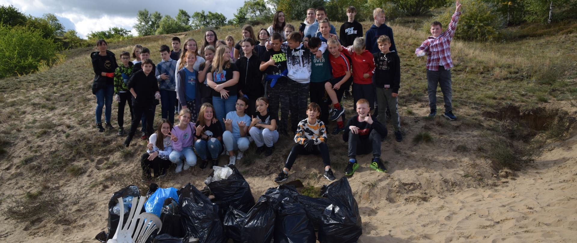 Na lekkim zboczu Gorzowskich Muraw pozują do zdjęcia uczestnicy sprzątania rezerwatu - grupa kilkudziesięciu osób dorosłych i dzieci ,. Za ich plecami zielone drzewa, krzewy. i trawy. . Poniżej leżą czarne worki z zebranymi odpadami.