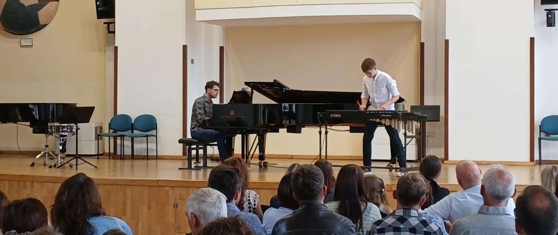 Na scenie sali koncertowej młody perkusista wykonuje utwór na marimbie, a towarzyszący mu pianista akompaniuje przy fortepianie koncertowym. Występ odbywa się przed licznie zgromadzoną publicznością siedzącą na widowni. Jasne wnętrze sali z wysokim sufitem i balkonem podkreśla uroczysty charakter koncertu.