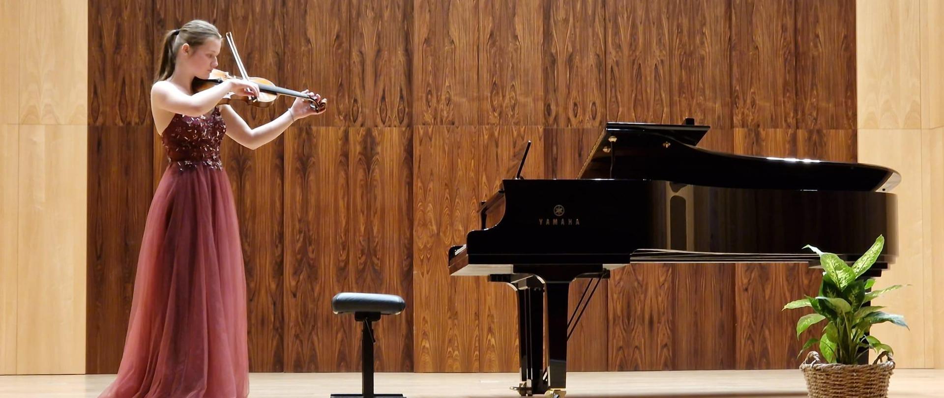 Uczennica na scenie gra na skrzypcach, w tle fortepian i kwiat w dniczce