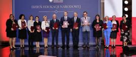 Pod niebieską ścianą z napisem Dzień Edukacji Narodowej stoi w szeregu grupa kilkunastu osób, trzymają czerwone teczki.