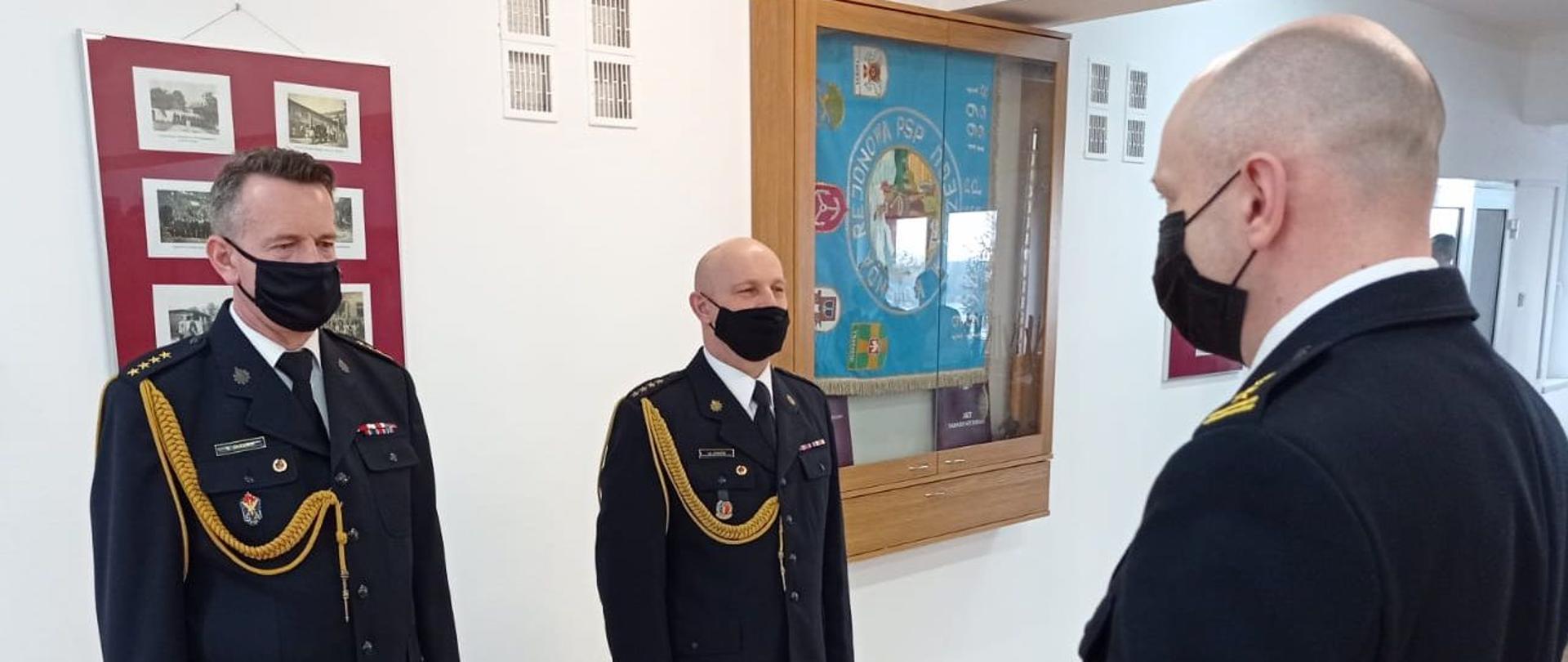 Na zdjęciu widoczni trzej strażacy w mundurach galowych z założonymi czarnymi maseczkami na twarzy. Po lewej stronie widoczny st. kpt. Sławomir Jakubik a 1,5 po lewej jego stronie stoi st. kpt. Mieczysław Ciróg. Twarzą do nich skierowany jest (plecami do fotografującego) bryg. Dariusz Gieroń Komendant Powiatowy PSP w Brzegu trzymający w dłoniach tabliczkę pamiątkową dla jednego z funkcjonariuszy.