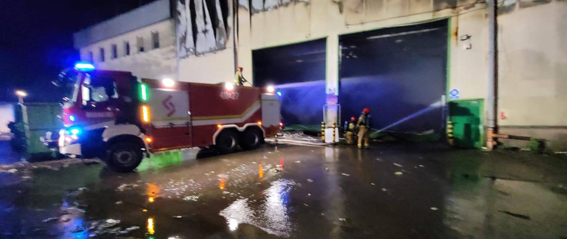 Zdjęcie zrobione nocą. Zdjęcie trochę rozmazane. Duży samochód strażacki stoi przy budynku i przez bramy wjazdowe podaje wodę przez działko dachowe do środka pomieszczenia. Trwa akcja gaśnicza.