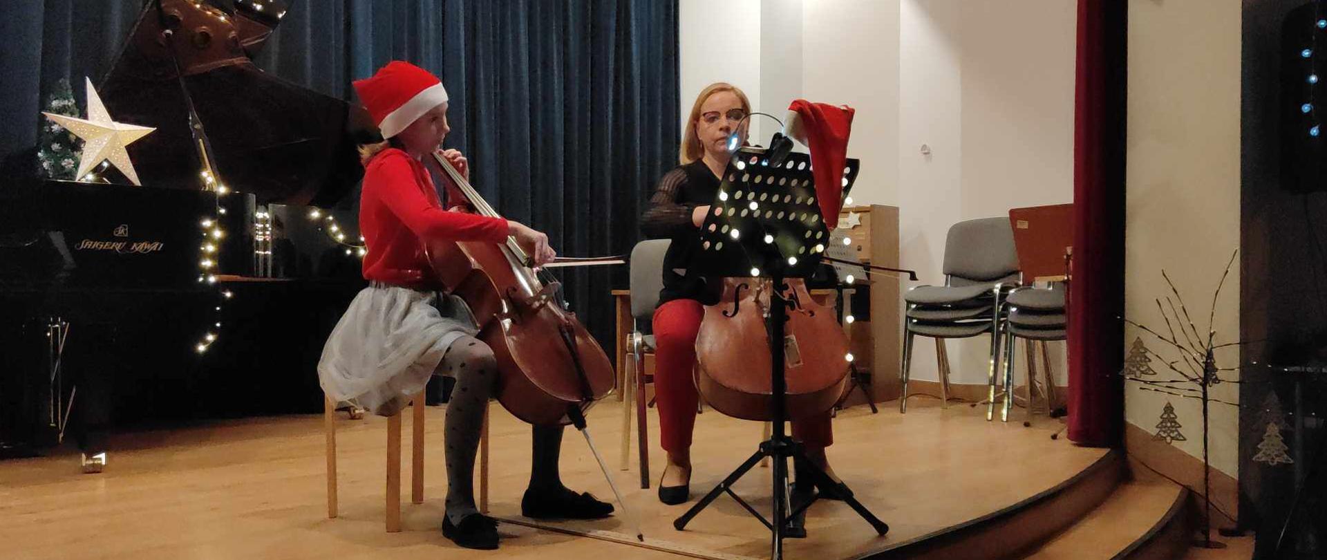Na świątecznie przystrojonej scenie znajdują się uczennica i nauczycielka, grające na wiolonczelach.