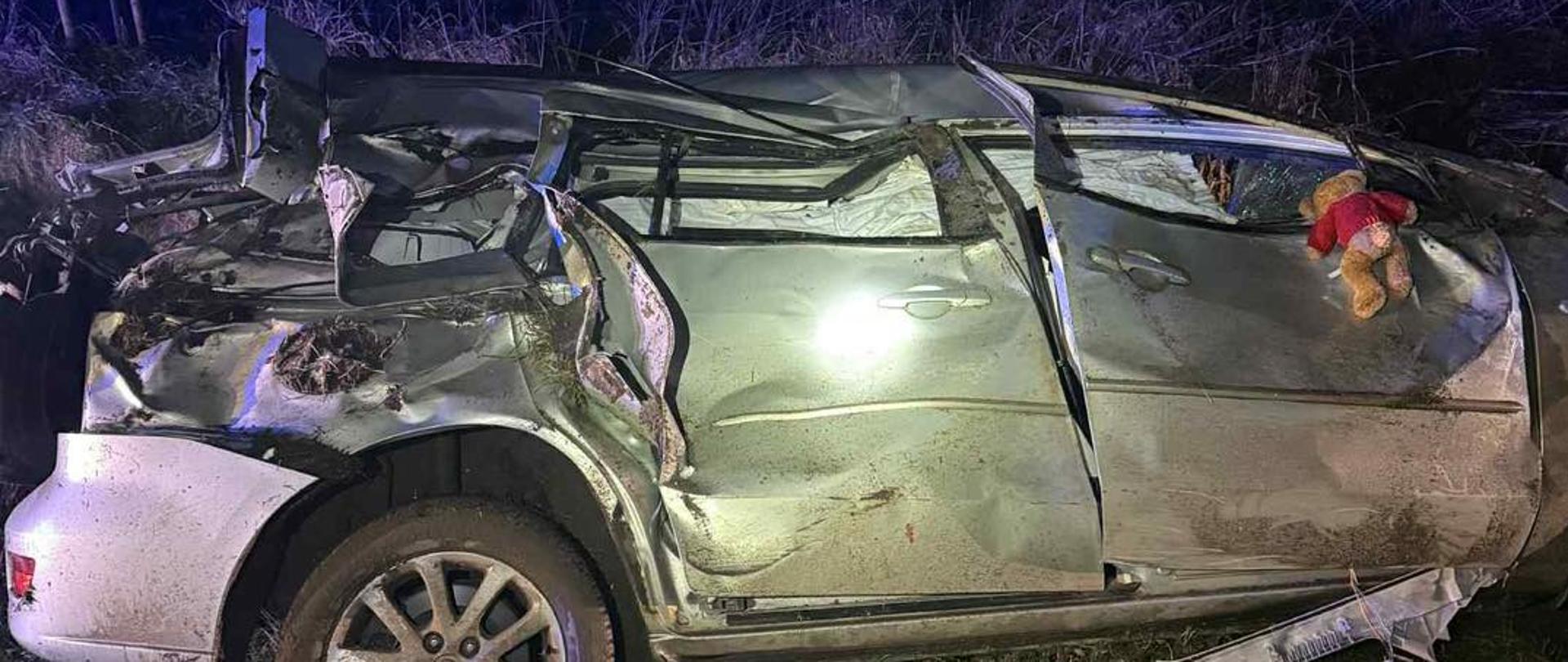Widok z boku na mocno zdeformowany samochód po ciężkim wypadku. Dach jest zapadnięty, a karoseria głęboko wgnieciona na całej długości pojazdu. Auto znajduje się na poboczu nocą, otoczone suchą roślinnością.