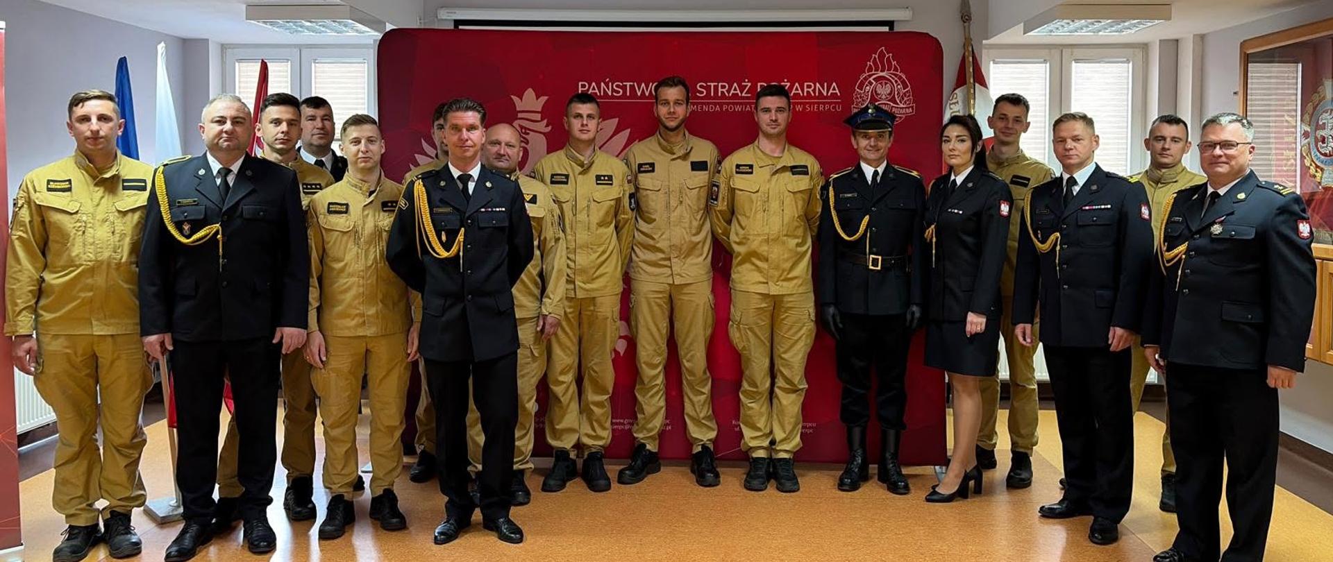 Group zbiorowe zdjęcie strażaków z Komendy Powiatowej Państwowej Straży Pożarnej w Sierpcu stojących w sali konferencyjnej na tle czerwonej ścianki prasowej.