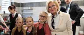 Trzy uśmiechnięte dziewczynki obok których stoi wiceminister Marzena Machałek ubrana w biały żakiet, białą bluzkę i apaszkę. W tle dziewczyna w kamizelce i krawacie 