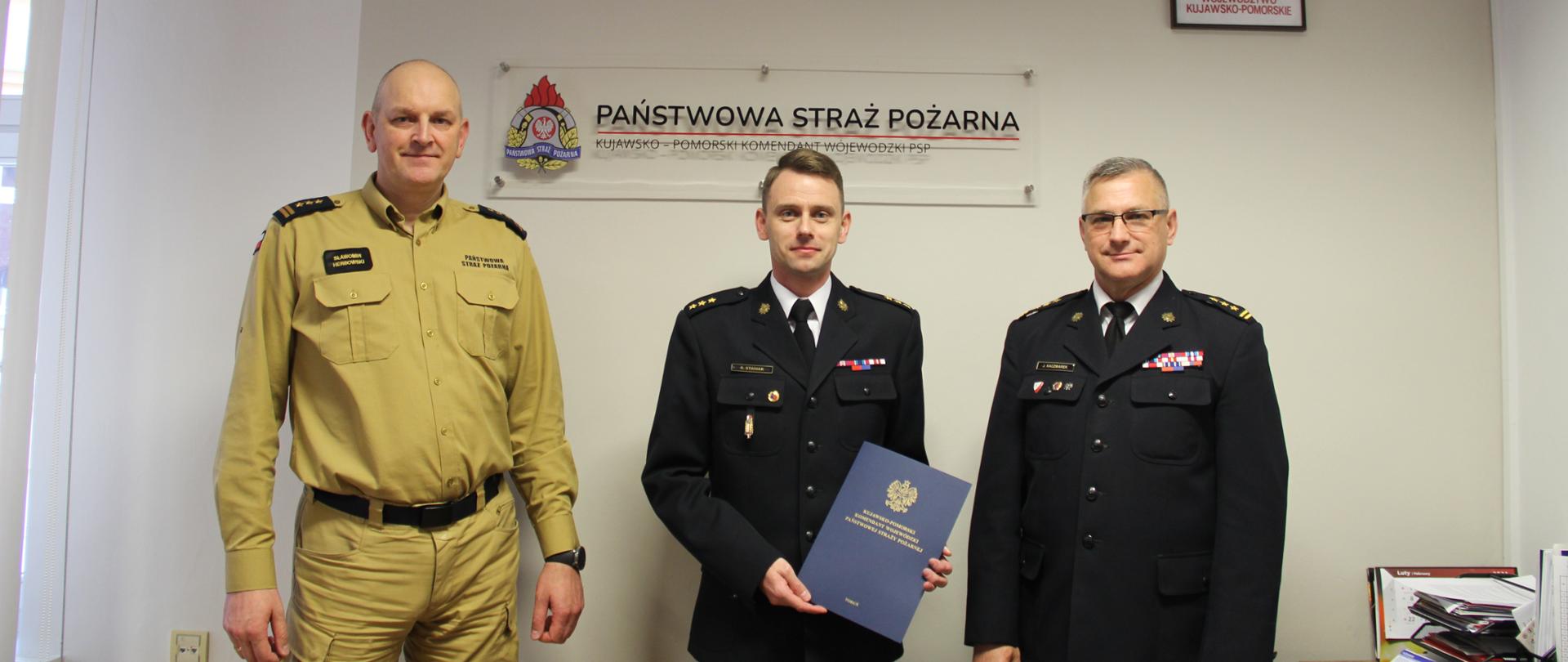 Na zdjęciu stoją od lewej: zastępca kujawsko kujawsko-pomorskiego komendanta wojewódzkiego PSP st. bryg. Sławomir Herbowski p.o. zastępcy komendanta powiatowego PSP w Wąbrzeźnie kpt. Robert Stasiak oraz kujawsko-pomorski komendant wojewódzki PSP st. bryg. Jacek Kaczmarek. Strażacy stoją na tle ściany nas nimi napis Państwowa Straż Pożarna, godło Polski oraz herb województwa kujawsko-pomorskiego. Nowo powołany zastępca trzyma w ręku akt powołania.