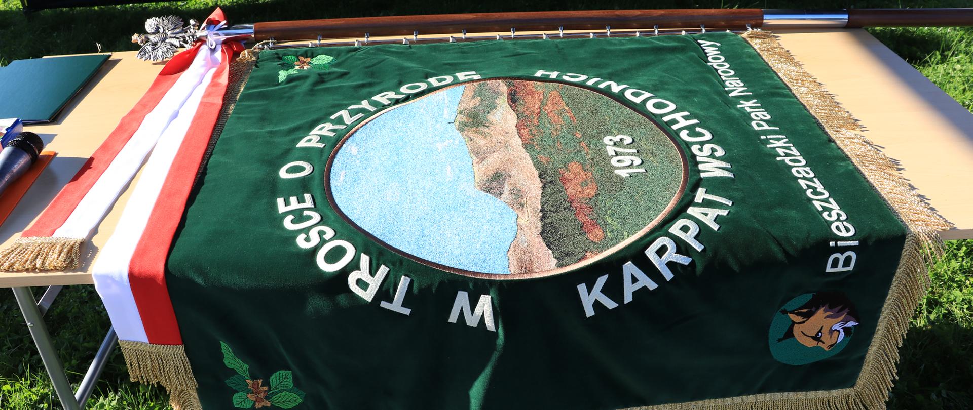 Na zdjęciu widać sztandar Bieszczadzkiego Parku Narodowego ułożony na stole. Jest On koloru zielonego z napisem w około panoramy gór bieszczadzkich "w trosce o przyrodę Karpat Wschodnich"