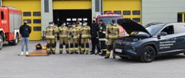 Zdjęcie przedstawia strażaków w ubraniu bojowym, stojących przed strażnicą przy samochodzie o napędzie hybrydowym