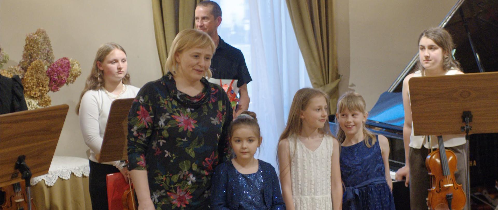W sali koncertowej podczas uroczystości pozuje do zdjęcia nauczycielka wraz z grupą dzieci i młodzieży. Część uczestników trzyma skrzypce, a w tle widoczny jest fortepian, pulpity nutowe oraz zasłonięte okna. Dzieci ubrane są elegancko, w jasne i granatowe stroje, a atmosfera wydarzenia ma uroczysty charakter.