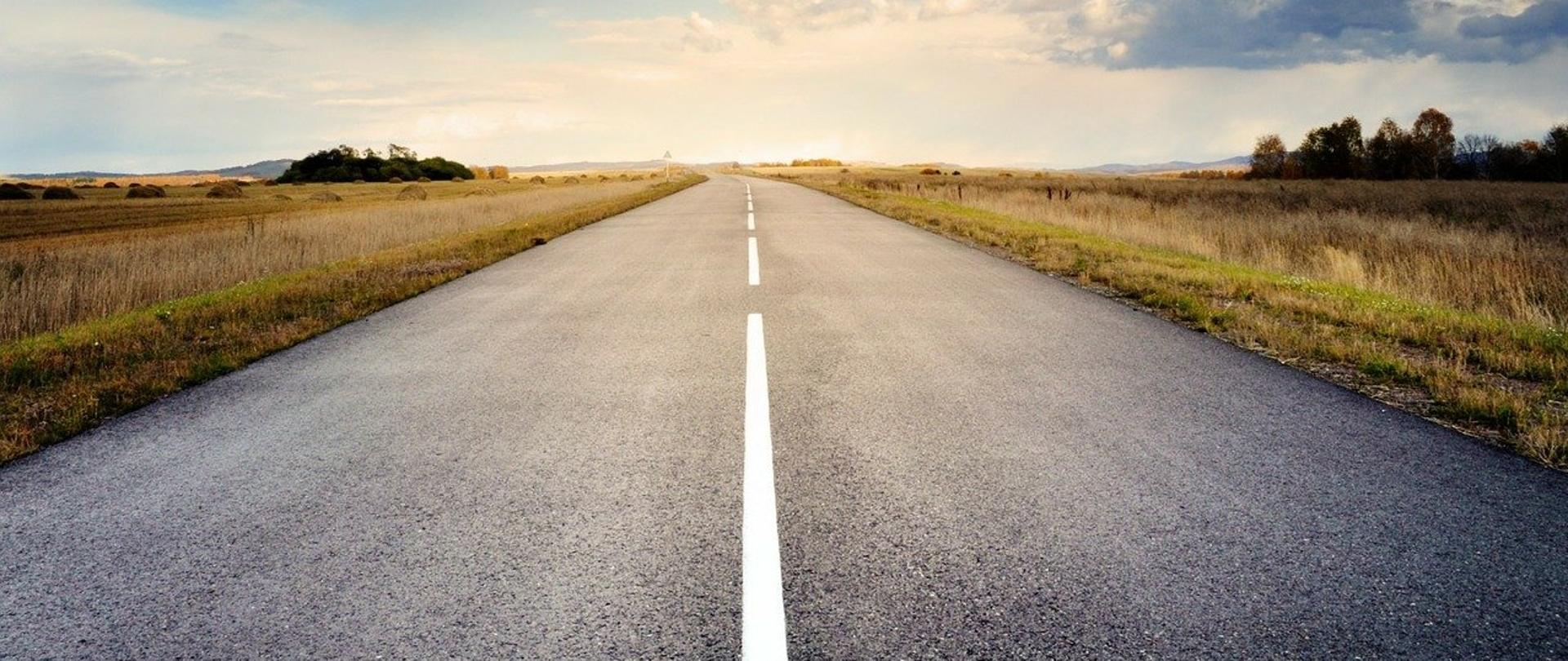 Zdjęcie polnej drogi, wybetonowanej. W środku kadru przebiegają linie oddzielające dwa pasy ruchu. W tle krajobraz. 