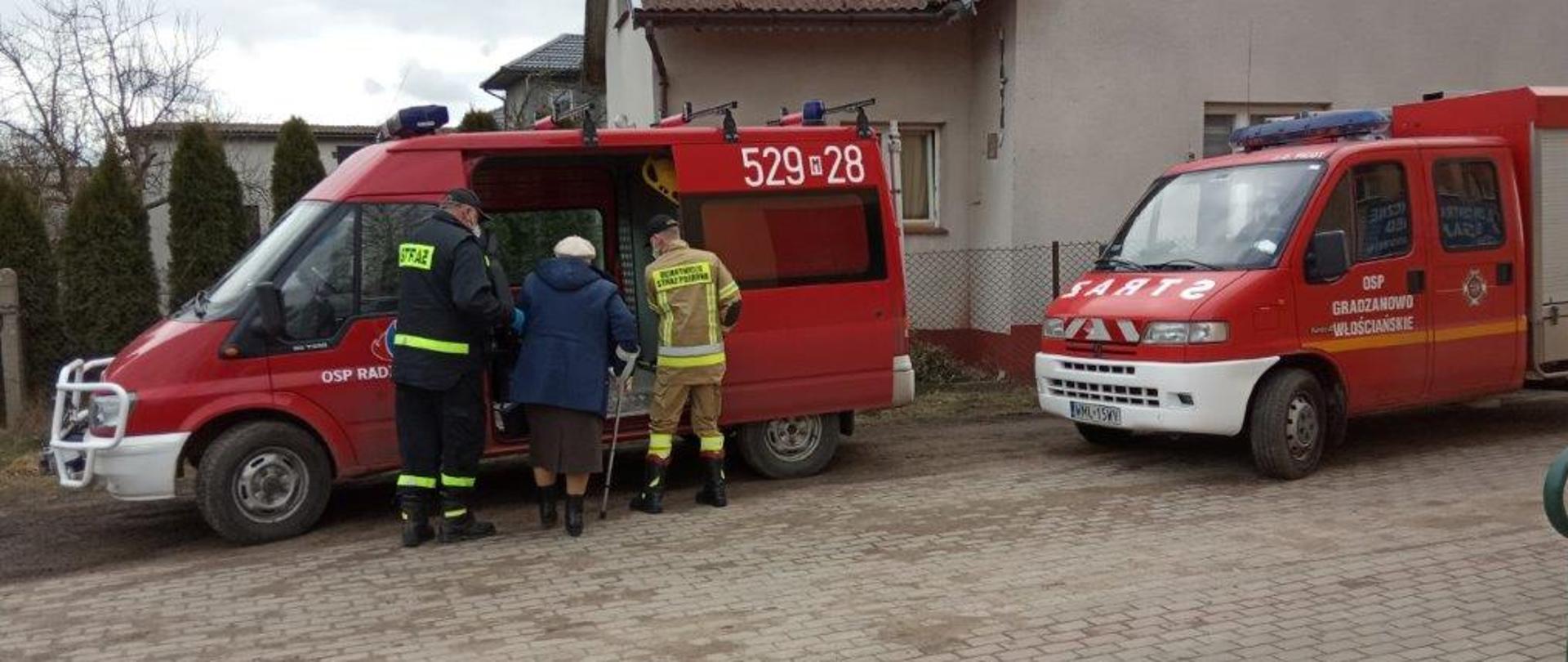 Na zdjęciu strażacy pomagają wejść seniorce do samochodu pożarniczego.