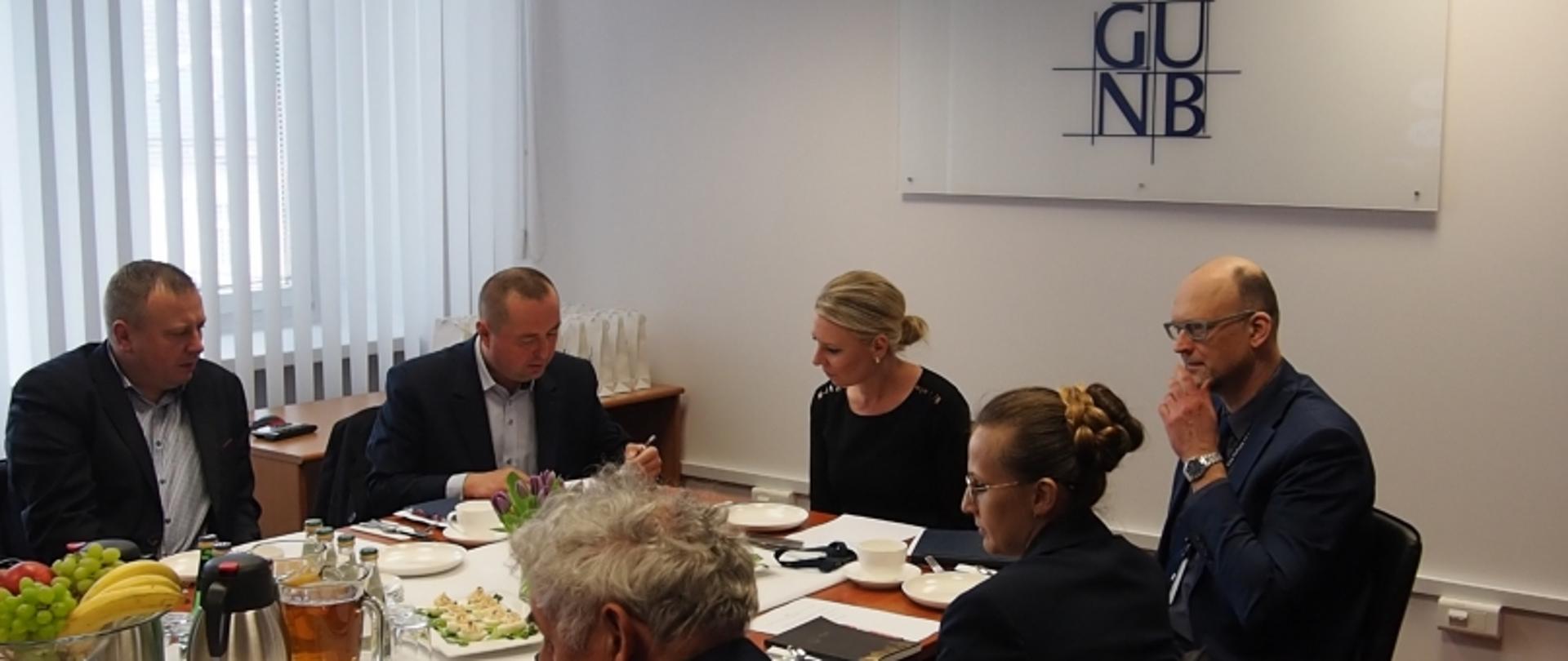 Grupa sześciu osób przy stole konferencyjnym z jedzeniem, napojami i dokumentami; w tle baner z napisem „GUNB”.