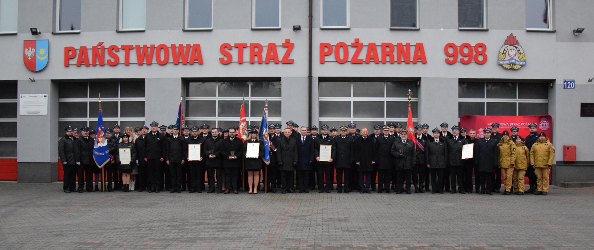 Goście uroczystości pozują do wspólnego zdjęcia na parkingu przed budynkiem z czerwonym napisem „PAŃSTWOWA STRAŻ POŻARNA 998”. 