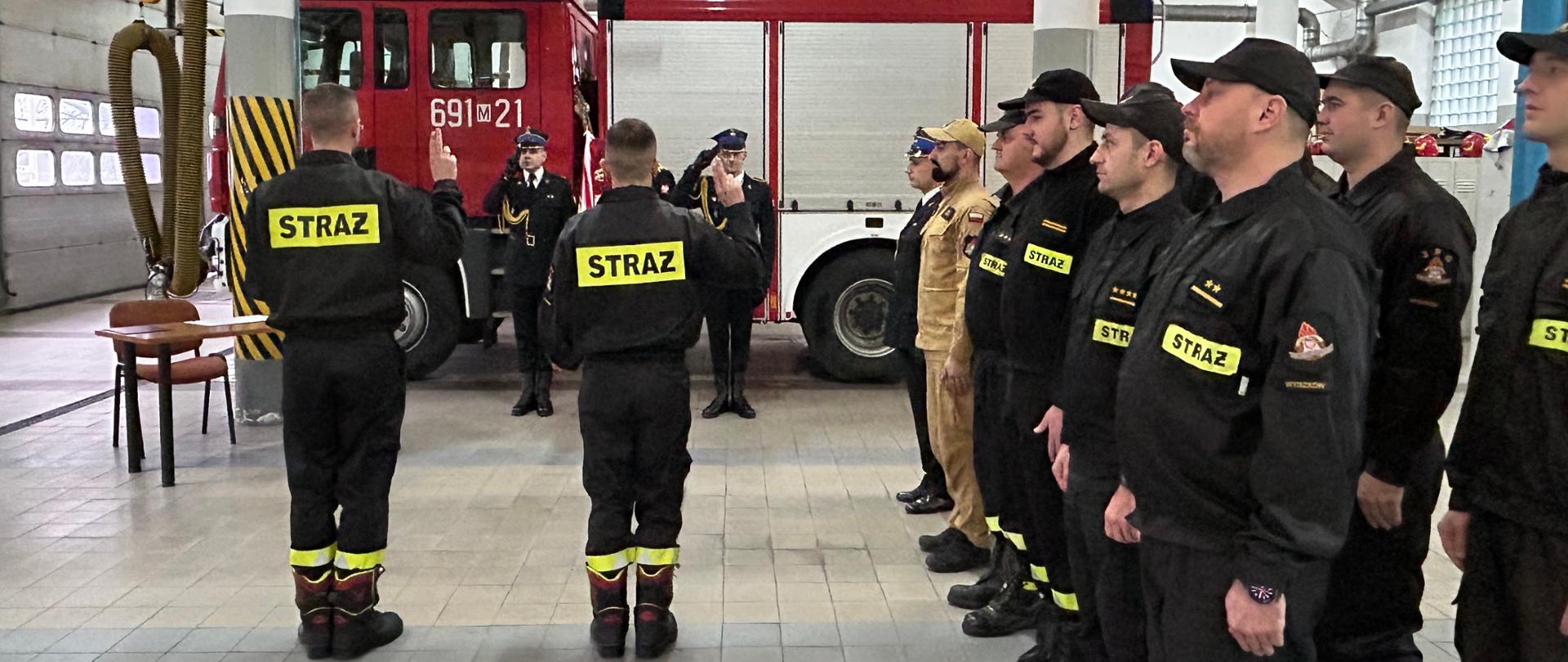 Strażacy składają ślubowanie przed sztandarem. Obok stoi dwuszereg strażaków.