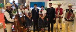 Wiceminister Marcin Ociepa w pawilonie polskim z zespołem ubranym w góralskie stroje regionalne.
