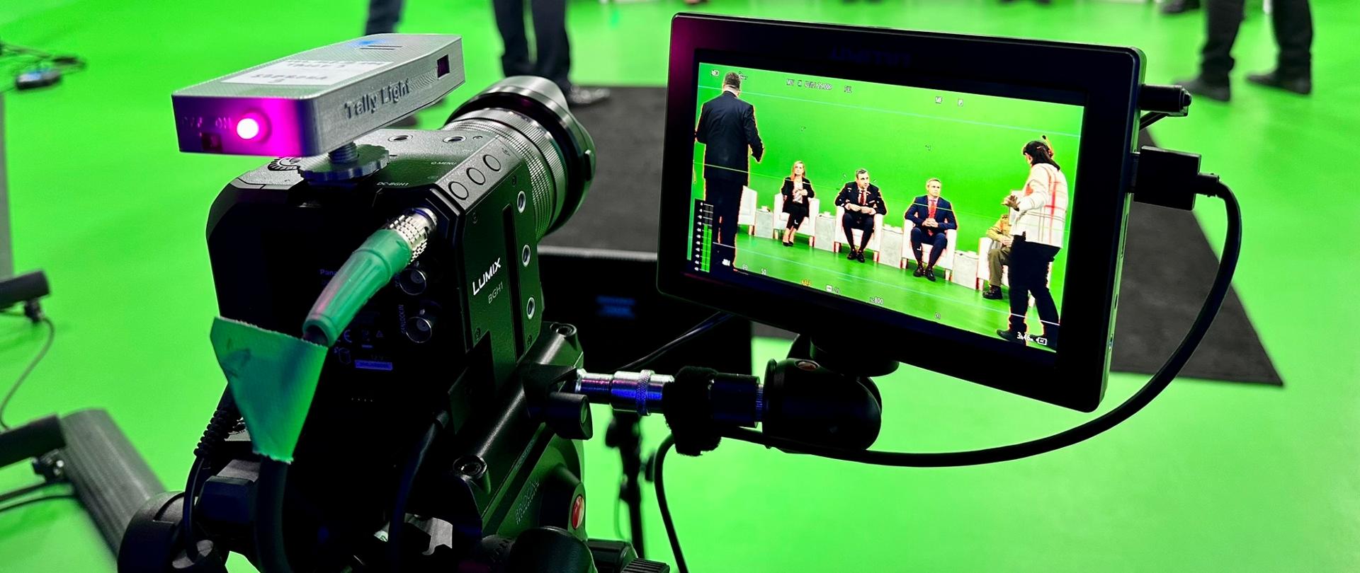 Zbliżenie na ekran kamery, a na nim cztery osoby, które siedzą na fotelach i dwie osoby stojące obok. 