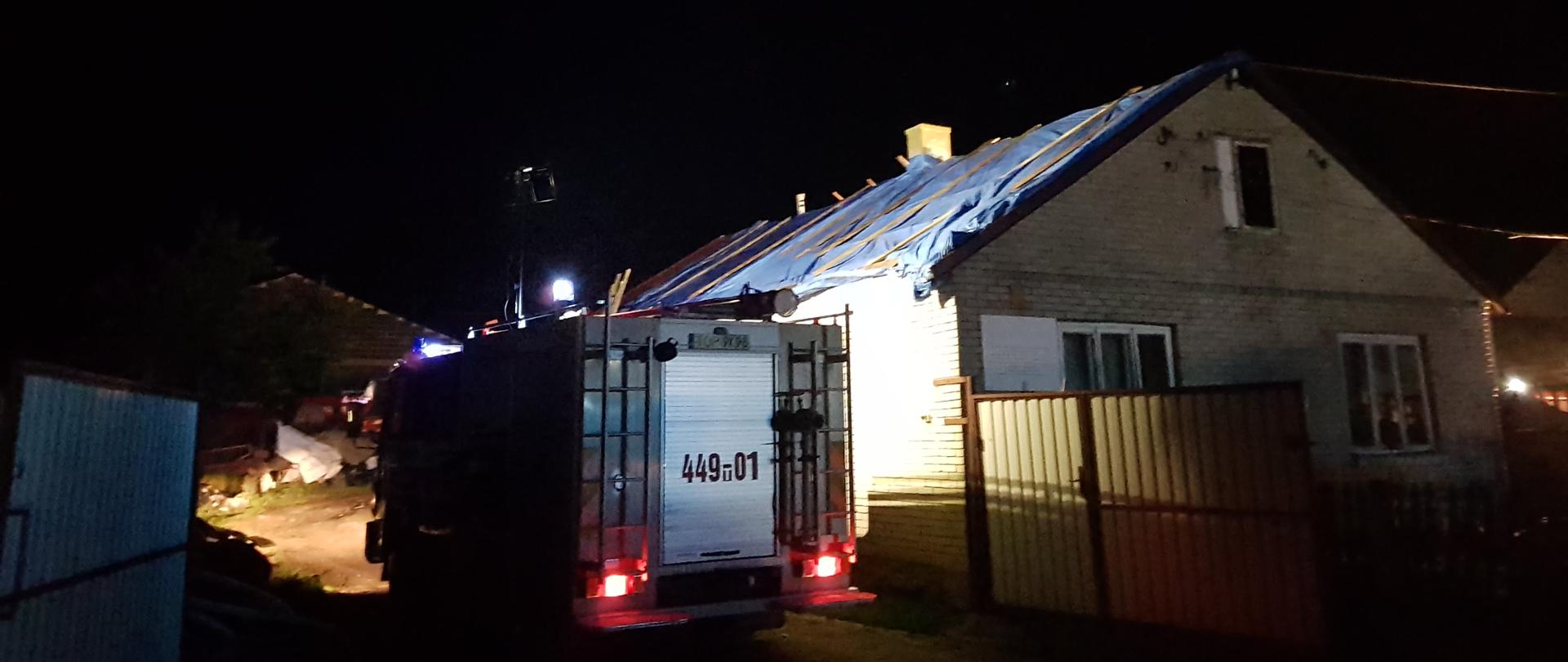Samochód gaśniczy stojący w bramie posesji prywatnej oświetlający budynek mieszkalny z dachem przykrytym niebieską plandeką przymocowaną deskami do więźby dachowej. W oddali pozostałości po zerwanym pokryciu dachowym.