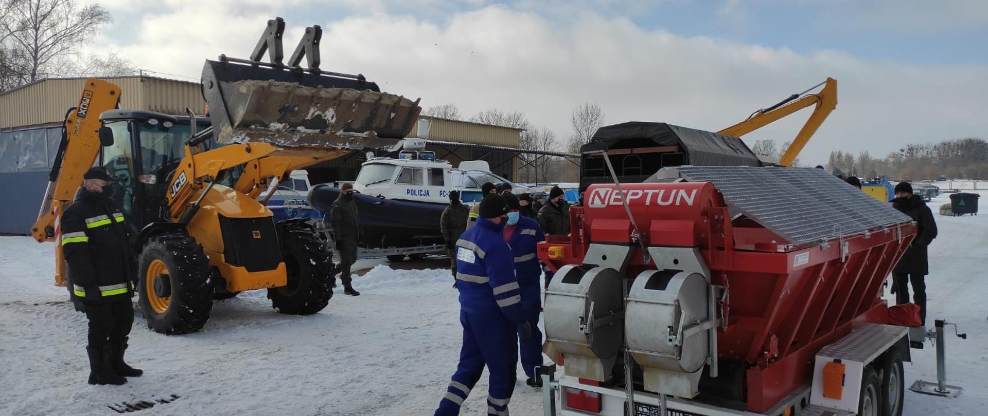 Na zdjęciu ciągnik z ładowaczem do piasku, na potrzeby urządzenia do wypełniania worków. Sprawdzenie stanu magazynu przeciwpowodziowego przed ewentualną powodzią. Na miejscu strażacy, żołnierze i przedstawiciele wojewody. 