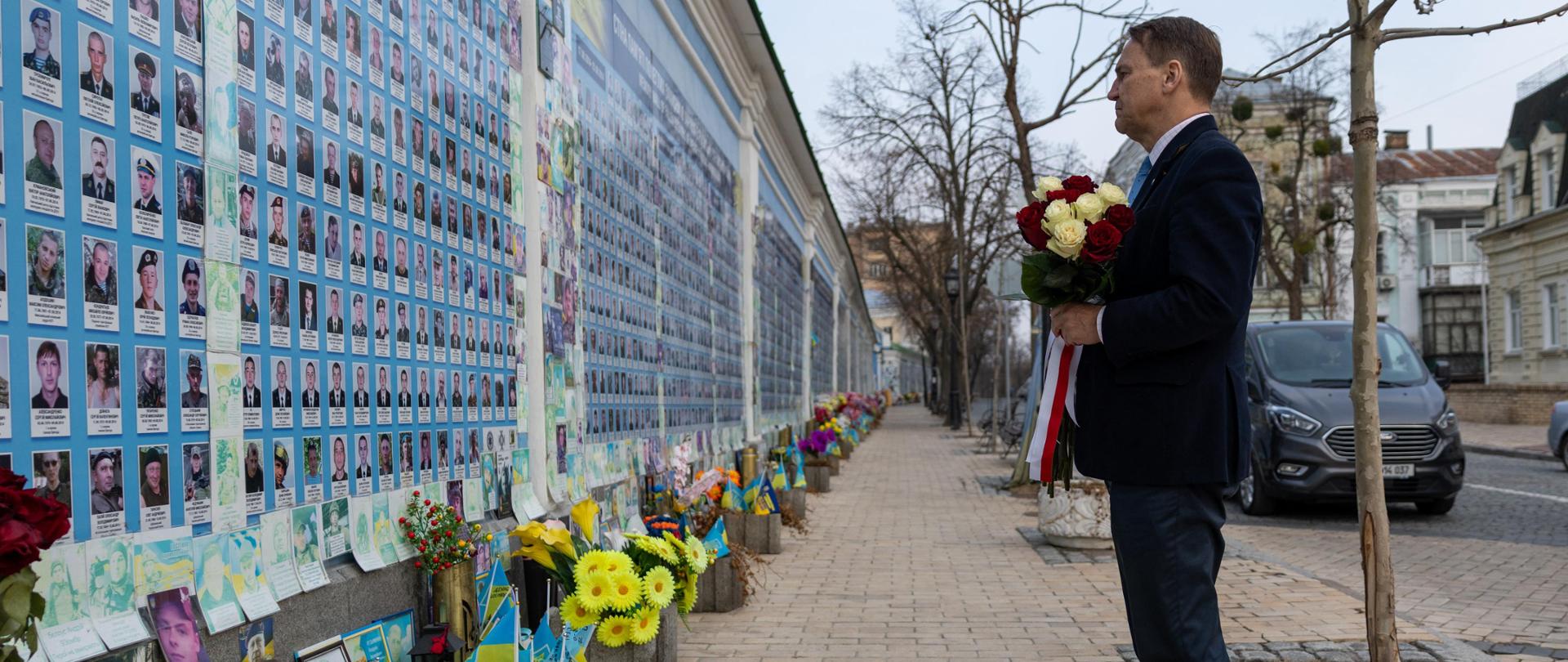 The photo shows a man in a suit. He is holding a bouquet of white and red roses. He is facing a wall covered with photos of many people. There are flowers at the foot of the wall. 