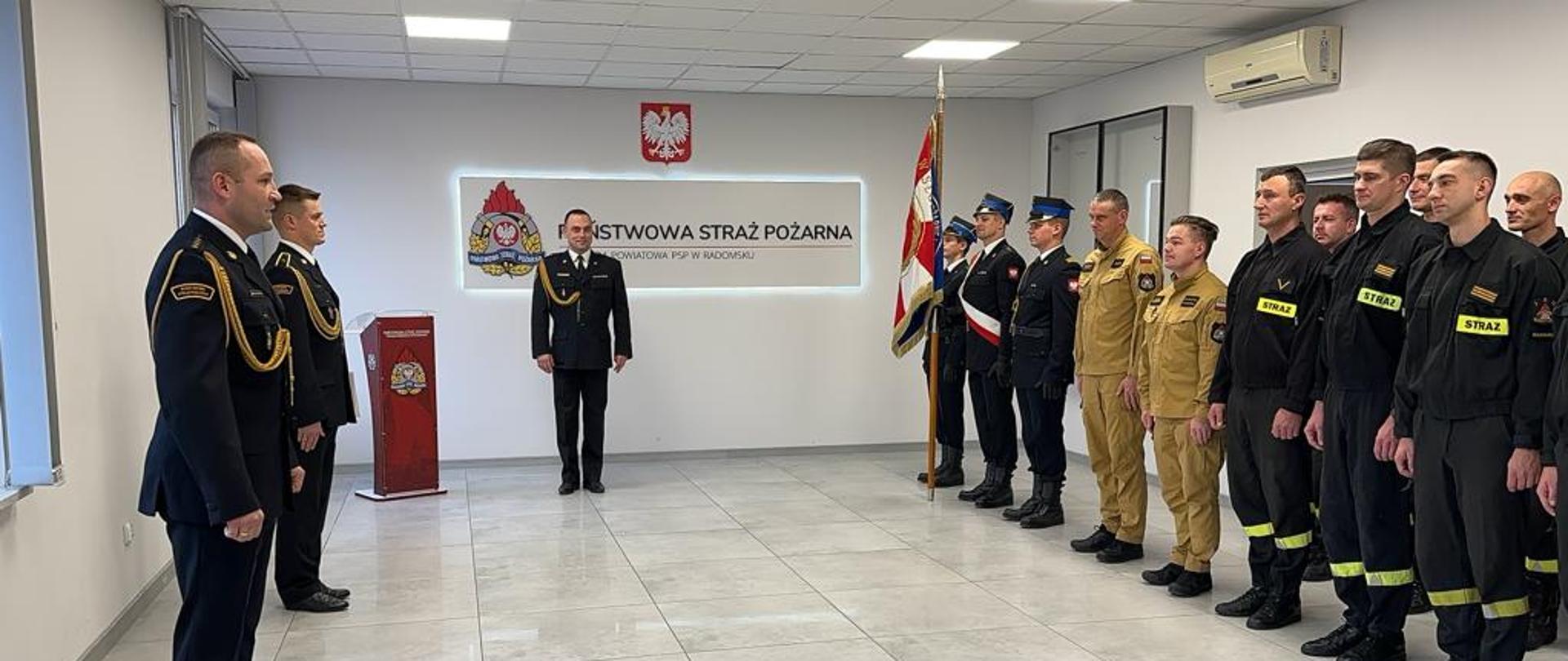 Zbiórka strażaków na uroczystości. Na środku sali Komendant. Po prawej stronie sztandar Komendy za nim rząd strażaków. W tle logo KP PSP w Radomsku oraz godło Rzeczypospolitej Polskiej.