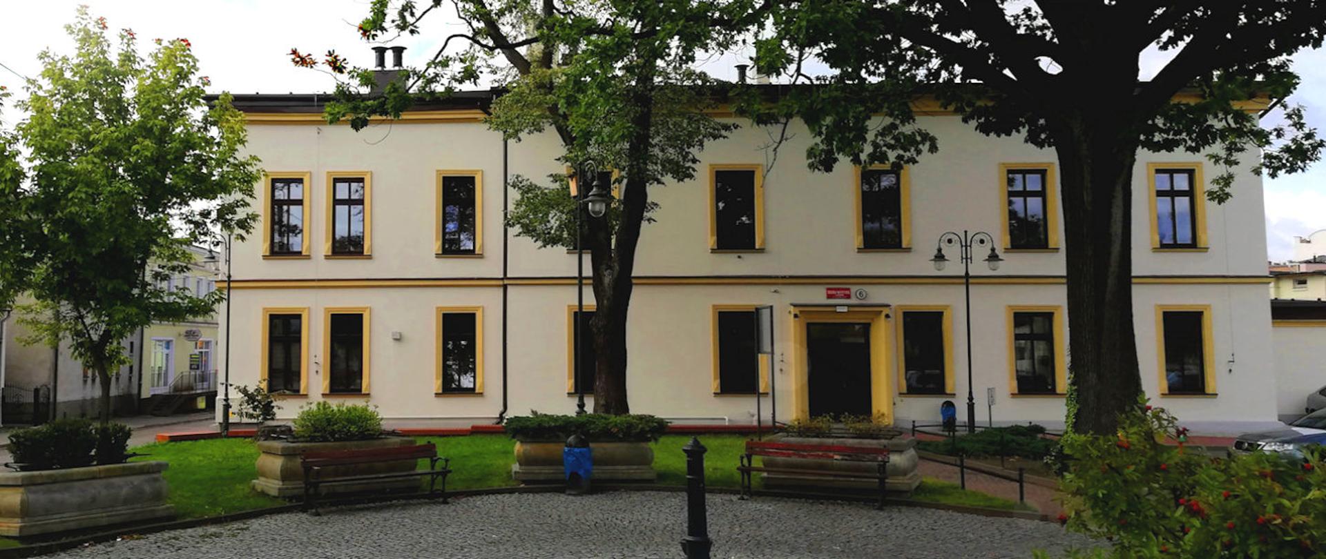 Zdjęcie przedstawia widok Szkoły Muzycznej I stopnia w Chojnicach od strony ul. Bankowej. Na zdjęciu widzimy front budynku szkolnego otoczony zielenią i małym placem z ławkami.
