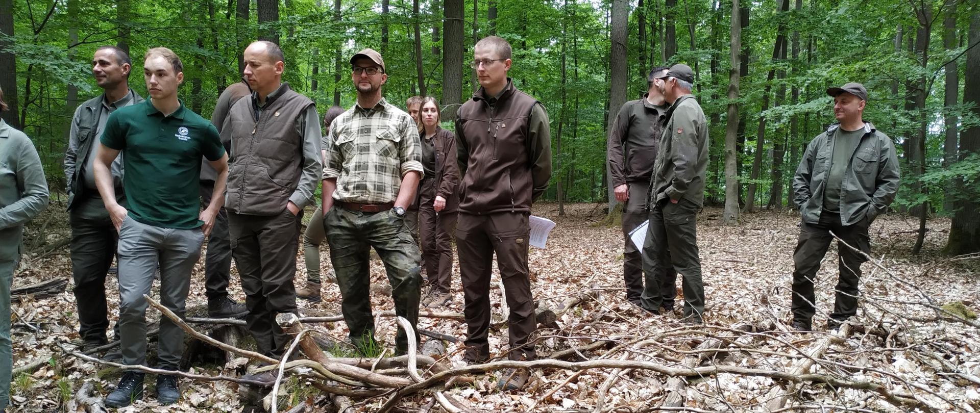 Grupa kilkunastu osób ubranych w zielone i brązowe stroje stoi w lesie. Za ich plecami zielone drzewa liściaste. Na ścieżce leżą suche, brązowe liście oraz gałęzie.