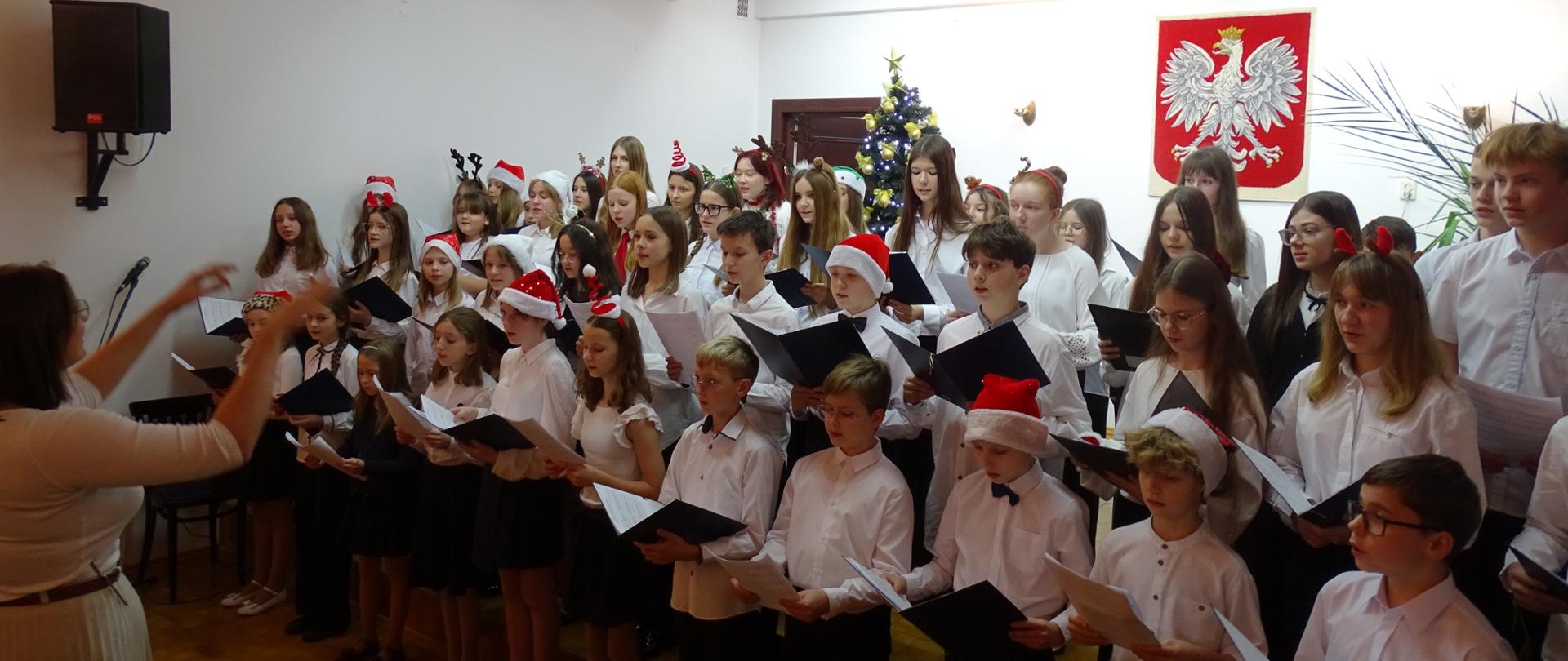 W sali koncertowej stoi w kilku rzędach grupa dzieci ubranych na galowo, niektórzy z nich mają na głowie czapki świętego mikołaja. W rękach trzymają nuty i śpiewają. Przed nimi stoi pani nauczycielka od chóru z uniesionymi rękami. W tle widać choinkę, a na ścianie godło Polski