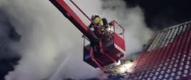 Zdjęcie przedstawia palący się dach w budynku dwurodzinnym przy którym stoi podnośnik koszowy strażacki z którego podawana jest woda. Zdjęcie zrobione nocą.