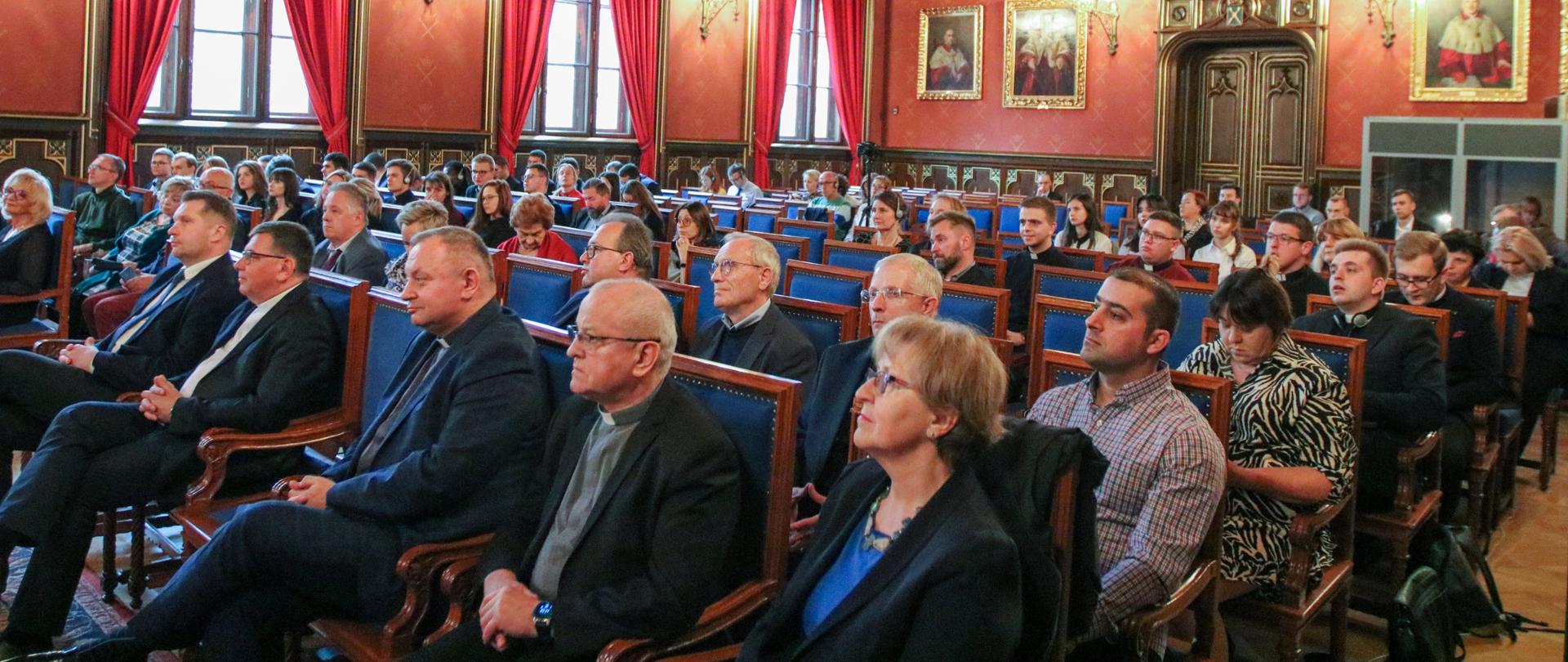 Grupa osób siedzi na konferencji w sali z portretami 