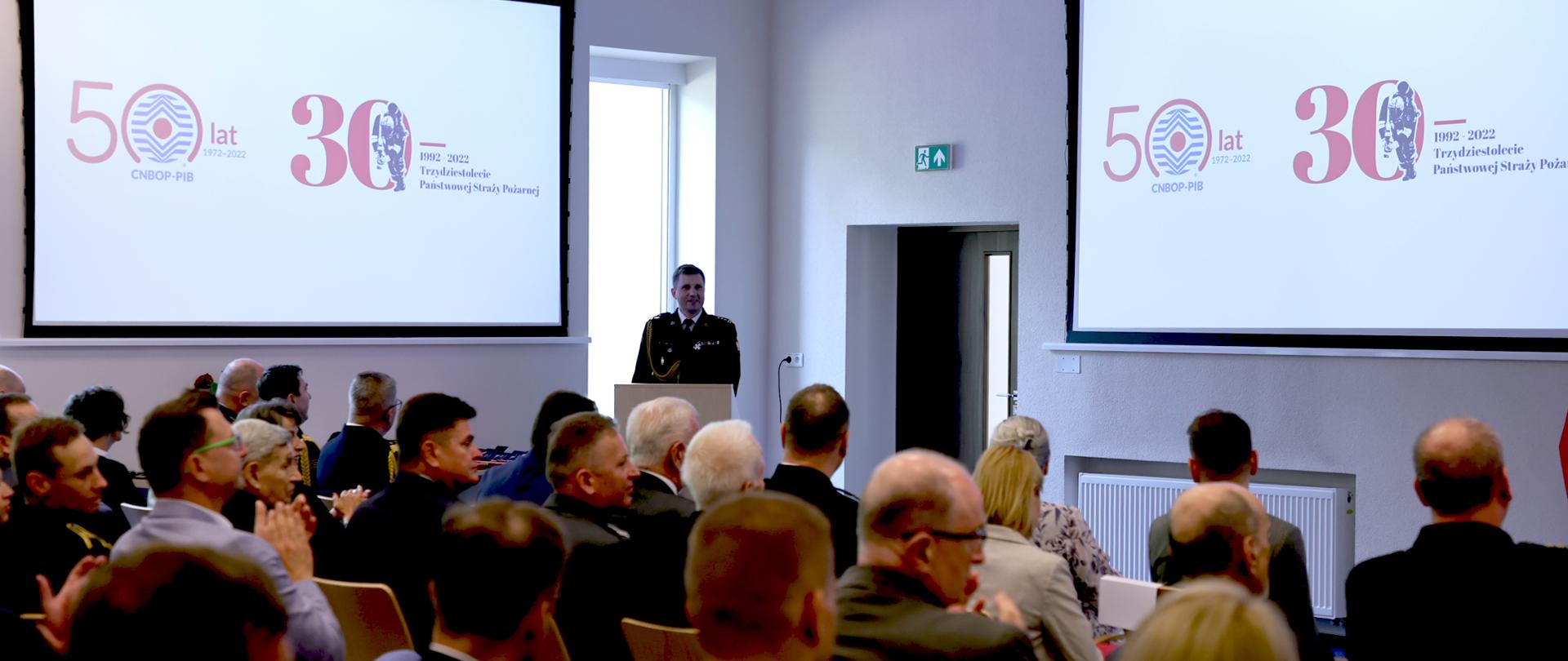 Widok ogólny sali na której odbywa się uroczystości. W tle mężczyzna przy mównicy oraz ekrany gdzie wyświetlane są logotypy 50lecia Instytutu oraz 30lecia PSP