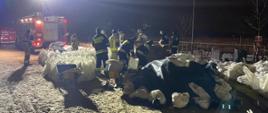 Strażacy napełniający worki z piaskiem podczas zabezpieczania wałów. Pora nocna. W oddali samochód strażacki oświetlający teren