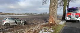 Zdjęcie przedstawia srebrny samochód osobowy w polu po zderzeniu z drzewem. Po prawej stronie znajduje sie wóz strażacki.