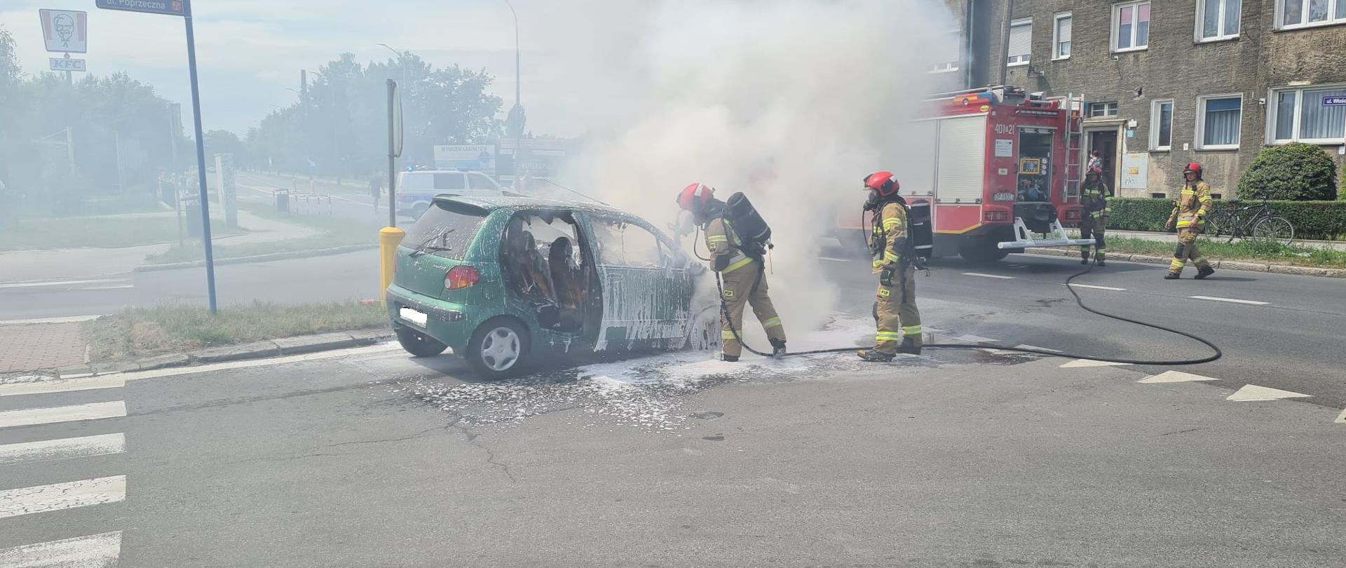 Pożar samochodu osobowego na skrzyżowaniu ul. Poprzecznej z ul. Chocimską w Brzegu - zdjęcie przedstawia strażaków ubranych w aparaty powietrzne podający prąd piany ciężkiej na palący się samochód osobowy w kolorze zielonym