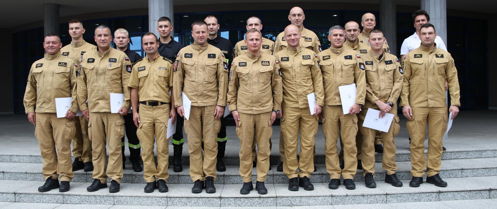 Pamiątkowe zdjęcie funkcjonariuszy PSP będących uczestnikami szkolenia (15 osób) w towarzystwie Zastępcy Komendanta Centralnej Szkoły PSP, mężczyzny oraz funkcjonariusza CS PSP