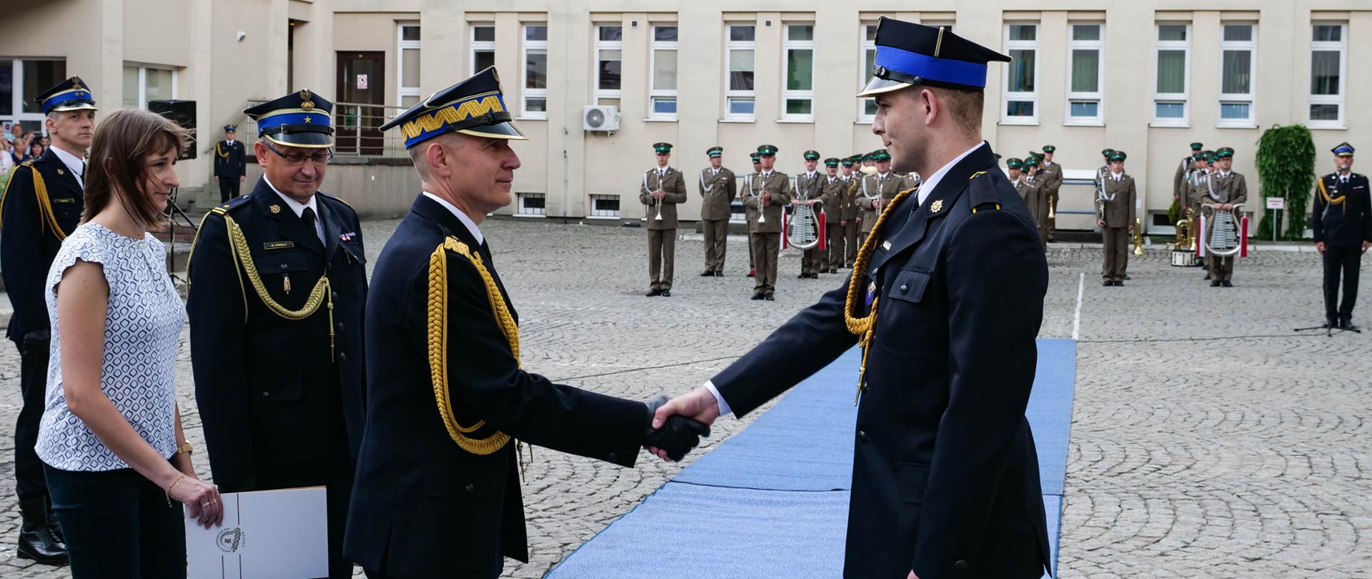 Komendant Główny PSP podaje rękę absolwentowi SA PSP, za Komendantem stoi kobieta trzymając w prawej dłoni białą teczkę papierową, po lewej stronie kobiety stoi Komendant Szkoły, w tle widoczna orkiestra w mundurach