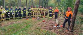 Strażacy oraz pilarze w ubraniach roboczych w lesie. Przed nimi leżą na trawie pilarki.
