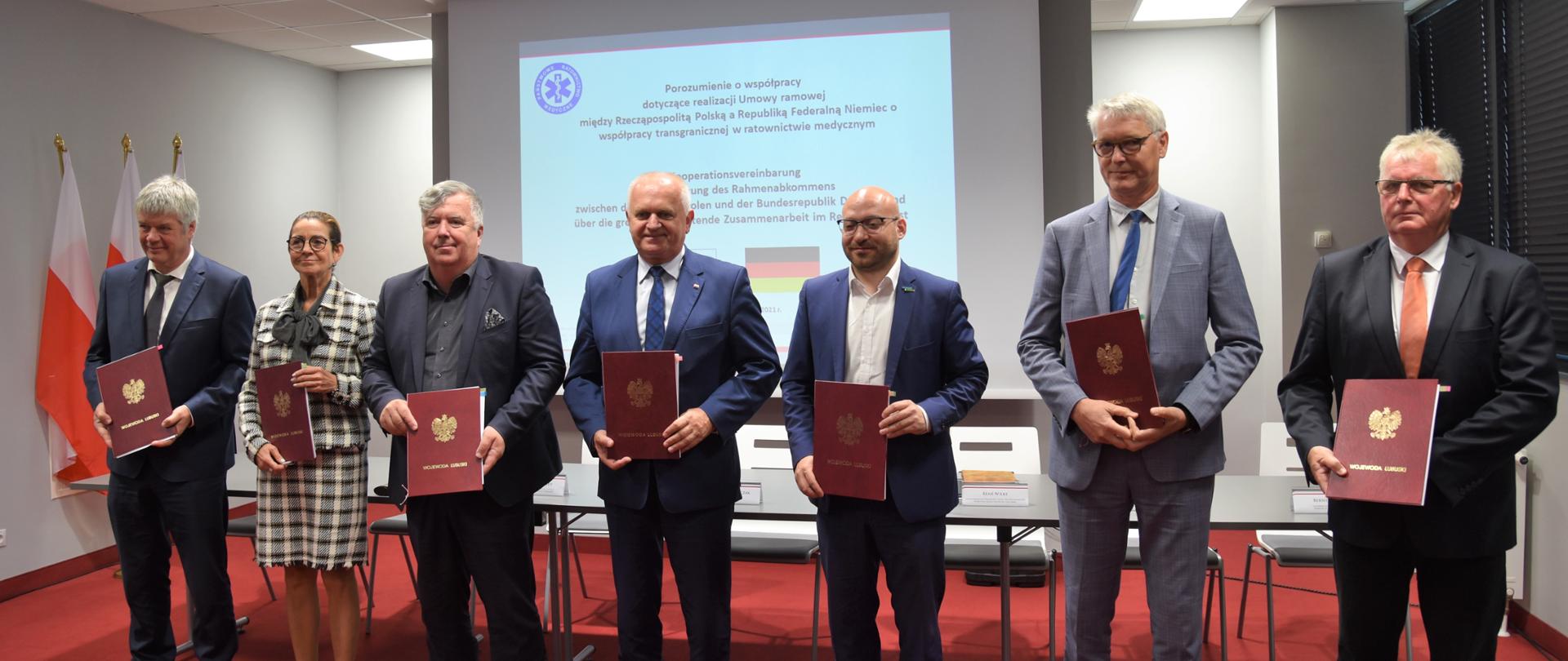 Sygnatariusze umowy stoją wspólnie, w jednym rzędzie, trzymają bordowe teczki z umowami 