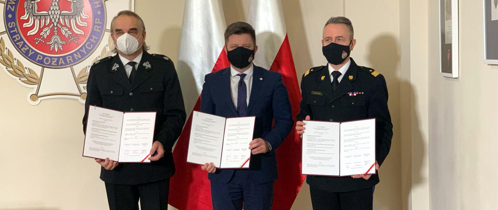 Zdjęcie przedstawia Michała Dworczyka, szefa Kancelarii Prezesa Rady Ministrów z nadbryg. Andrzejem Bartkowiakiem, komendantem głównym PSP i dh. Waldemarem Pawlakiem prezesem ZG ZOSP RP, przedstawiających podpisany list intencyjny w sprawie Narodowego Programu Szczepień przeciw COVID-19