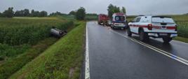 Wypadek drogowy - auta ratownicze stoją na jezdni a rozbite auto osobowe w przydrożnym rowie na dachu
