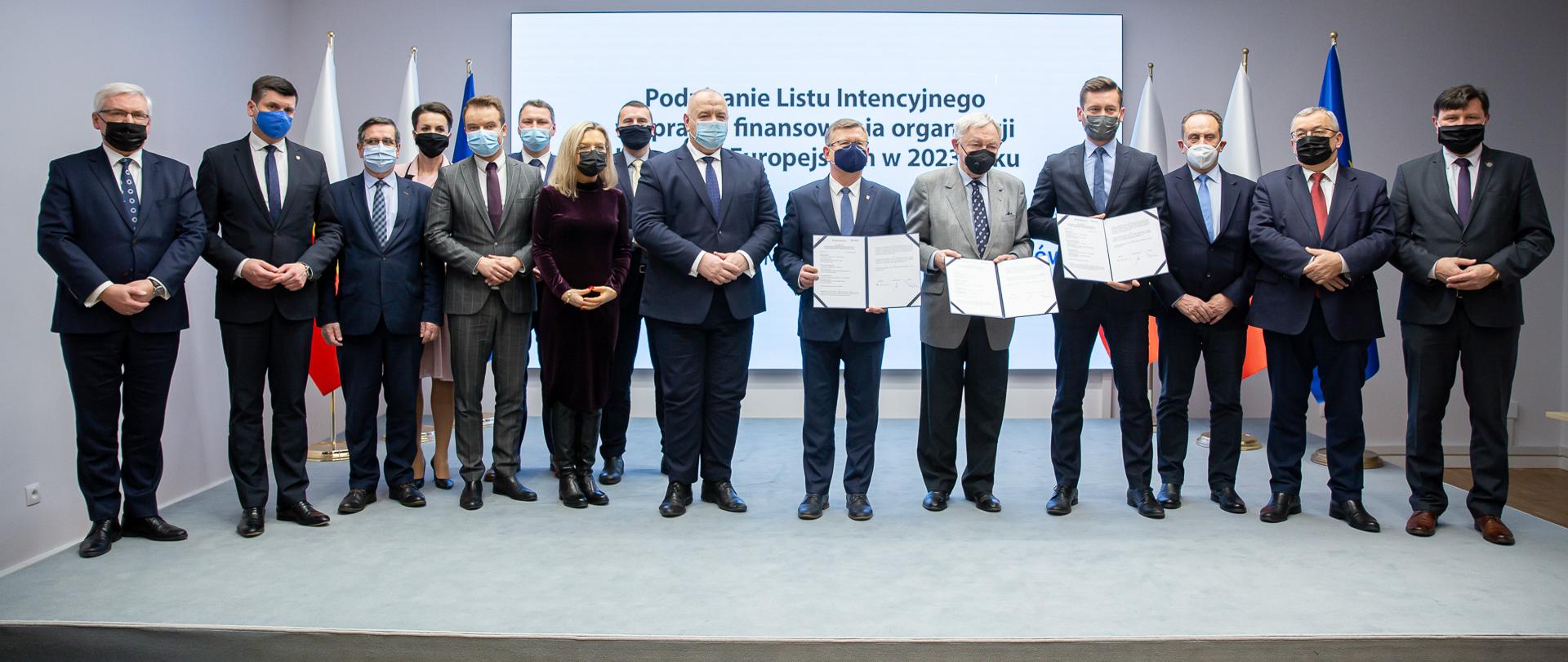 Zdjęcie grupowe uczestników uroczystości. Minister sportu, prezydent Krakowa i marszałek województwa trzymają w rękach otwarte teczki z listem intencyjnym. Na środku stoi wicepremier Jacek Sasin