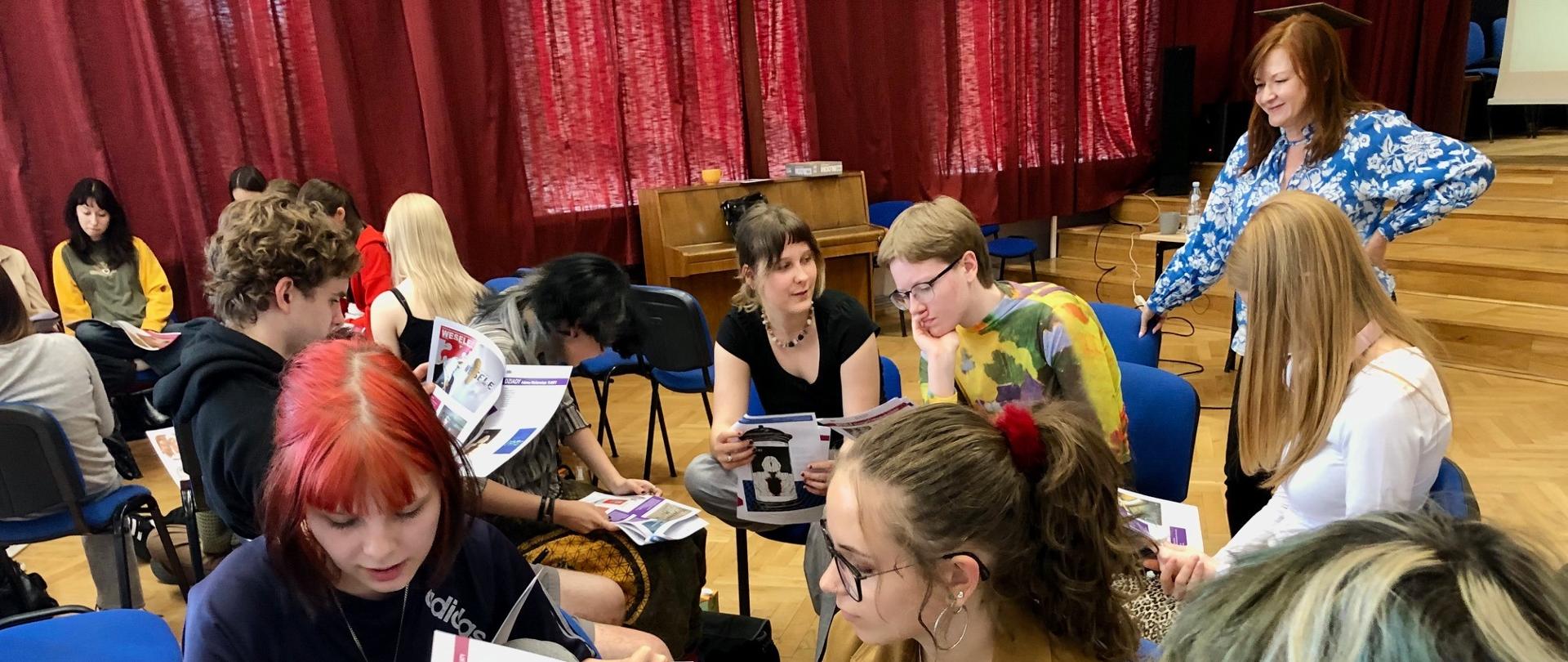 Grupa młodzieży siedzi w kręgu w szkolnej sali z drewnianą podłogą i czerwonymi zasłonami. Uczniowie przeglądają kolorowe materiały edukacyjne z napisem „Wesele”, co sugeruje, że uczestniczą w zajęciach dotyczących dramatu Stanisława Wyspiańskiego. W tle nauczycielka w niebieskiej koszuli z kwiatowym wzorem obserwuje grupę z uśmiechem. Atmosfera jest skupiona, ale zarazem swobodna.