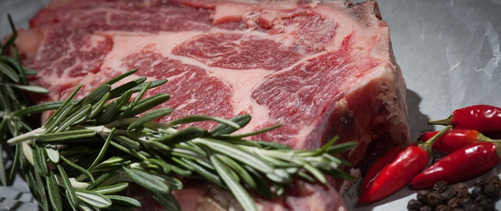 Kawałek surowego mięsa na blacie, na nim gałązka ziół, obok czerwona papryka oraz pieprz w ziarnach