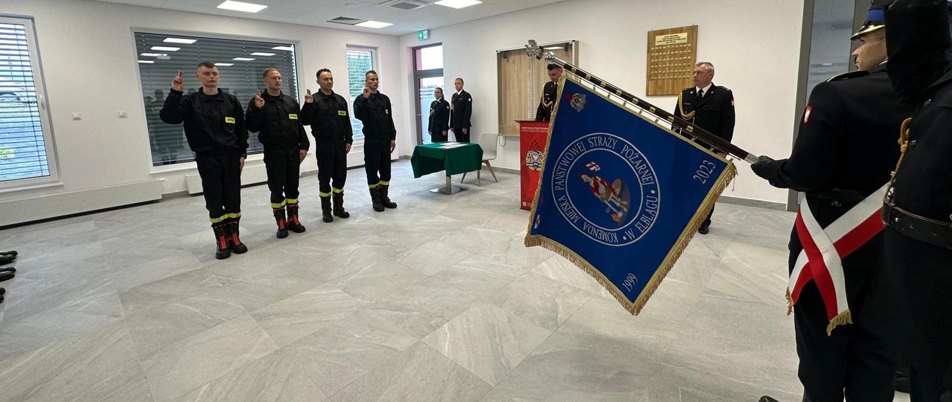 Na sali odpraw nowo przyjęci funkcjonariusze, w ubraniu koszarowym składają ślubowanie. Stoją na przeciwko pocztu sztandarowego. Mają uniesioną prawą dłoń i zgięte palce. W tle stoi kierownictwo komendy miejskiej. Funkcjonariusze mają na sobie mundury wyjściowe.