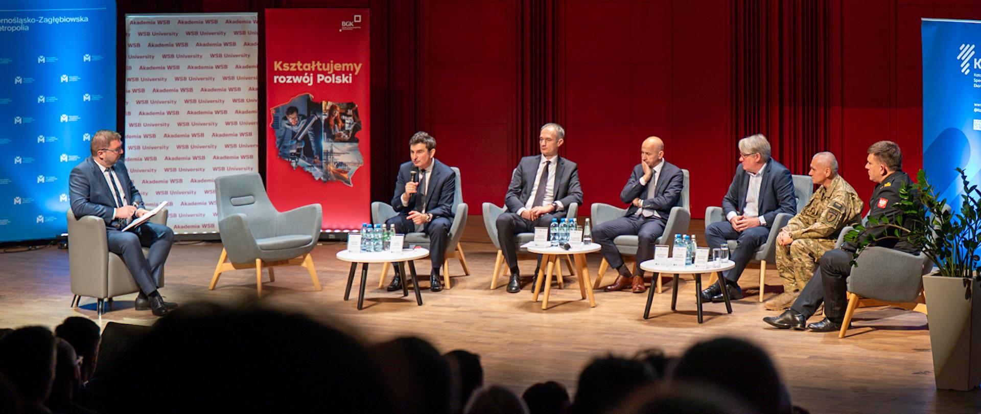 Na zdjęciu widać panel dyskusyjny odbywający się na scenie w sali konferencyjnej lub auli. Na scenie siedzi siedmiu mężczyzn – sześciu uczestników panelu i jeden prowadzący. Wszyscy mają mikrofony i są ubrani w garnitury, z wyjątkiem dwóch osób po prawej stronie: jeden z nich nosi mundur wojskowy, a drugi mundur strażacki. Przed uczestnikami znajdują się małe stoliki z butelkami wody i tabliczkami z nazwiskami. W tle widać czerwone panele ścienne i trzy pionowe banery: pośrodku biały i czerwony z logo „Akademia WSB” oraz napisem „Kształtujemy rozwój Polski”,
Na górze ekranu wyświetlony jest napis informujący o temacie konferencji: „Odporność cywilna i gospodarcza”, a także data i miejsce wydarzenia – Tychy, 27 października 2025 rok. Na pierwszym planie, w cieniu, widać sylwetki słuchaczy siedzących na widowni.