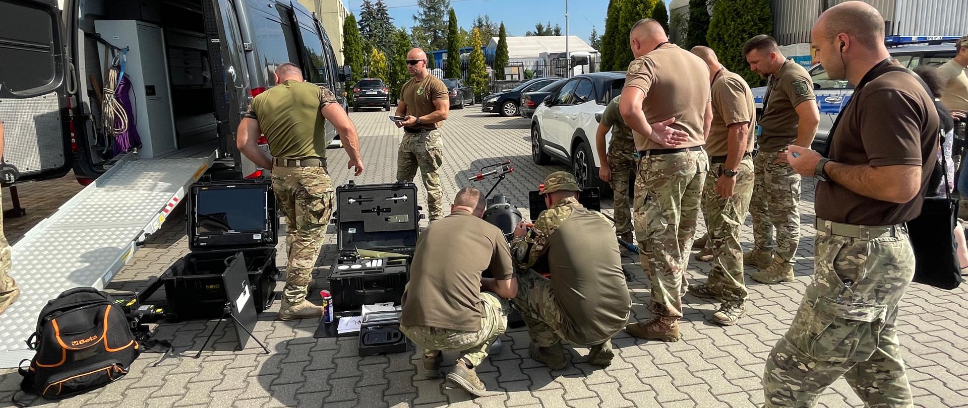 Na brukowanym parkingu obok półciężarówki grupa mężczyzn ogląda sprzęt umieszczony w kilku walizkach