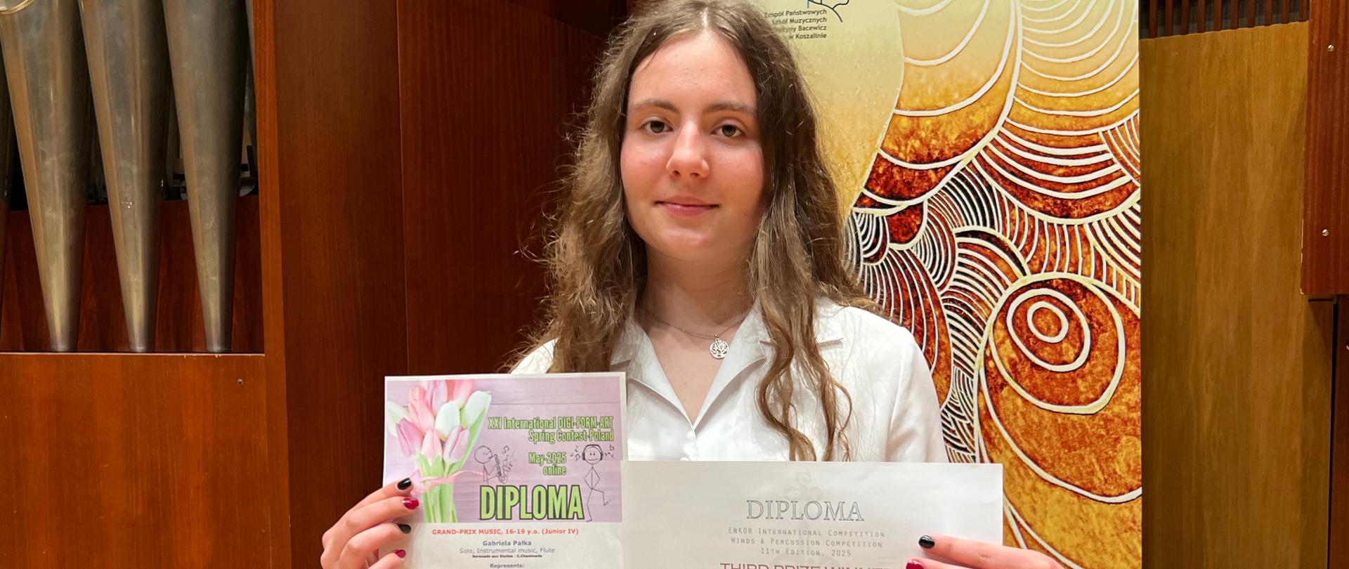 Uczennica trzyma dwa dyplomy, na tle banera ZPSM