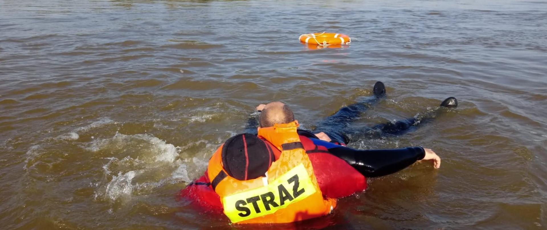 Zdjęcie przedstawia dwóch mężczyzn. jeden w kamizelce z napisem straż wydostaje drugiego na łódź. W oddali pomarańczowe koło ratunkowe a na trzecim planie kaja i tereny zielone