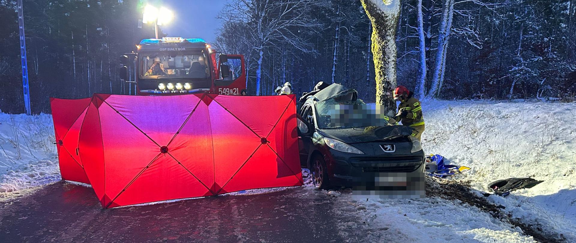 Akcja ratunkowa na oblodzonej drodze leśnej: rozbity samochód osobowy stoi przy drzewie, a strażacy pracują obok pojazdu. Przed nimi ustawiony jest czerwony parawan osłaniający miejsce zdarzenia, a w tle widać wóz strażacki z włączonymi światłami oraz śnieg pokrywający jezdnię i otaczający las.