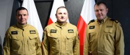 Zdjęcie zrobione wewnątrz pomieszczenia. Na zdjęciu trzech oficerów Państwowej Straży Pożarnej. Za nimi ustawione są biało-czerwone flagi.
